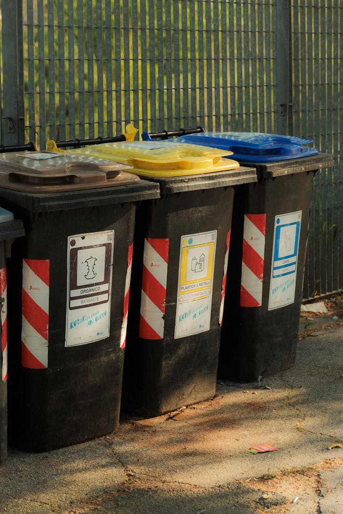 Four colorful recycling bins arranged outdoors next to a metal fence in sunlight.